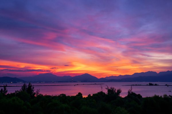 A breathtaking view of vibrant sunset hues reflecting over a calm lake surrounded by silhouetted mountains. Nature 4K