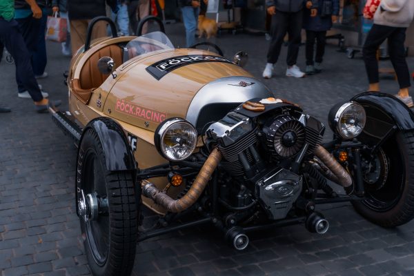 A classic three-wheeler car showcased outdoors, surrounded by people within a city environment. Voitures 4K