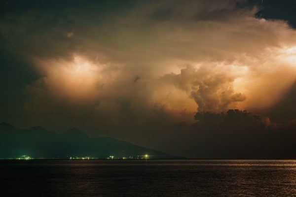A dramatic thunderstorm with vivid lightning illuminates clouds over a calm body of water at night. Dark 4K