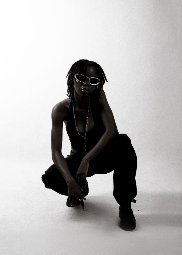 A fashionable young adult crouching, wearing sunglasses and a chain, in a minimalist studio setting. Mobile 4K