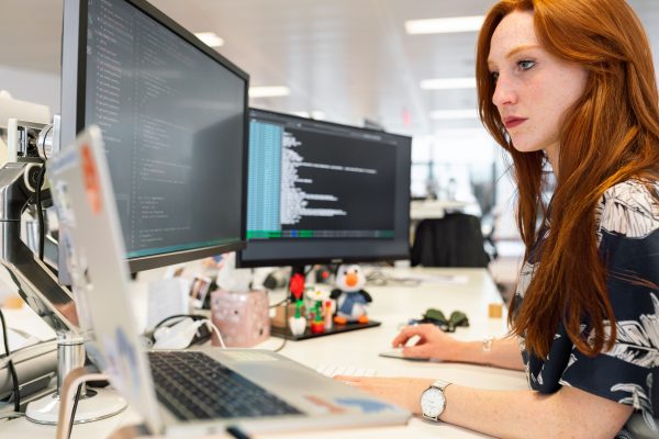 A focused female software engineer coding on dual monitors in a modern office. Wallpaper 4K