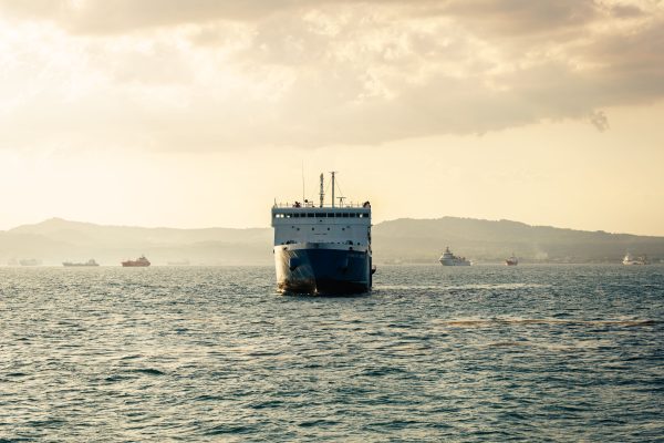 A large cargo ship sailing across calm waters with a golden sunset backdrop, surrounded by distant hills and ships. Nature 4K