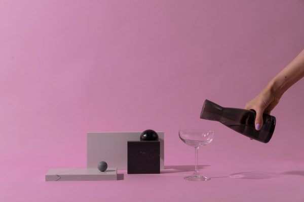A minimalist still life scene featuring a hand pouring from a pitcher over geometric objects on a pink surface. Minimaliste 4K