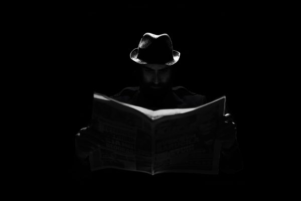 A mysterious figure in a fedora engrossed in reading a newspaper under dramatic lighting. Dark 4K