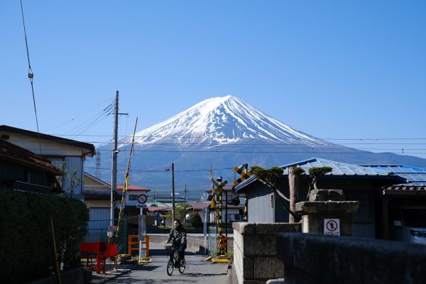 A person cycling near a railway crossing with Mount Fuji in the background under a clear blue sky. Anime 4K