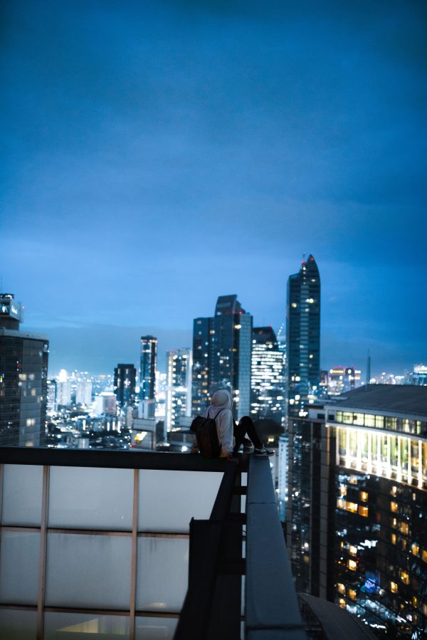 A person sits on a building's edge overlooking a city skyline illuminated at night. Mobile 4K