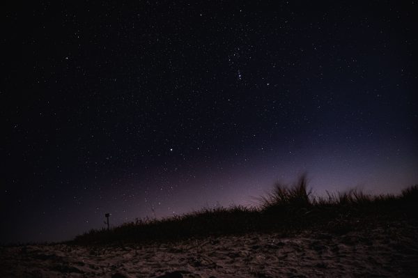 A serene view of stars twinkling over a calm beach with silhouetted sand dunes at night. Espace 4K