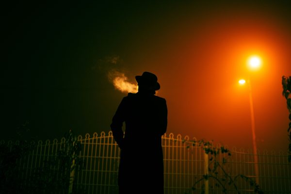 A silhouetted person in a hat smoking under an orange streetlight in a foggy night. Dark 4K