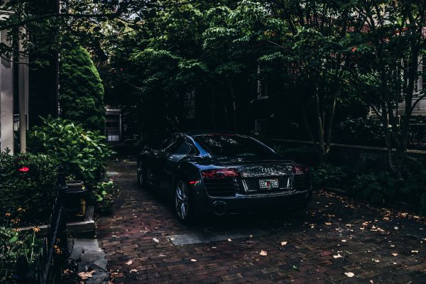 A sleek black Audi sportscar parked on a brick driveway surrounded by lush trees on a sunny day. Voitures 4K
