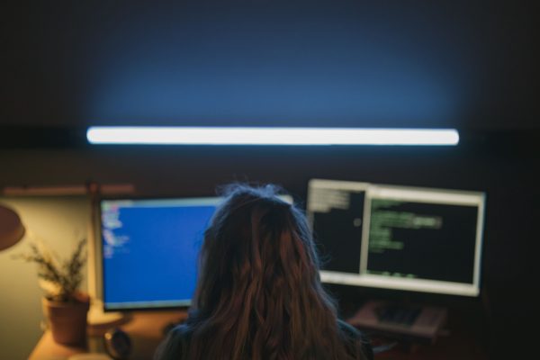 A software developer working in a dim-lit room with dual screens showing code, illuminated by a white light. Wallpaper 4K