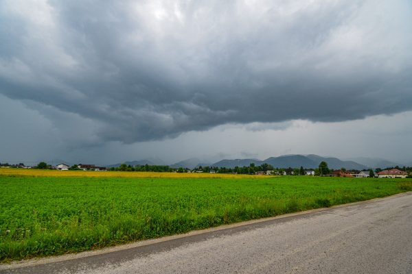 A storm brews over a lush green field with distant mountains and village houses. Dark 4K