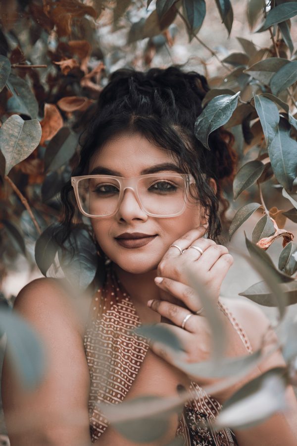 A thoughtful portrait of a woman with glasses surrounded by lush green leaves. Mobile 4K