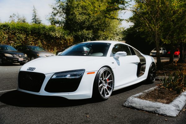 A white sports car parked in a sunny outdoor setting, surrounded by trees and vehicles. Voitures 4K