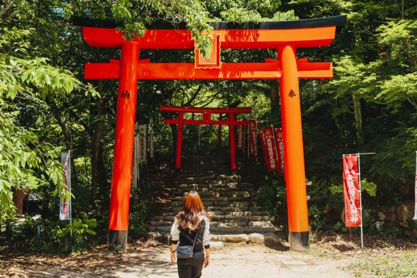 A woman approaches the vibrant red torii gates of Tenkai Inari Shrine in Fukuoka, Japan, surrounded by lush greenery. Anime 4K