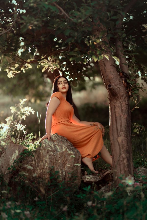 A woman in an orange dress sits on a rock under a tree, surrounded by greenery in Tehran, Iran. Mobile 4K