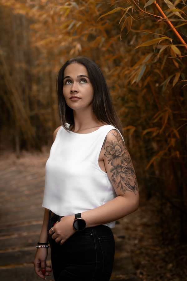A woman poses in a forest wearing a white tank top and black pants, showcasing tattoos. Mobile 4K