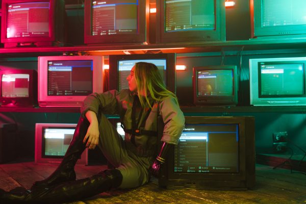 A woman with a prosthetic hand sits in a neon-lit room surrounded by retro TVs. Geek 4K
