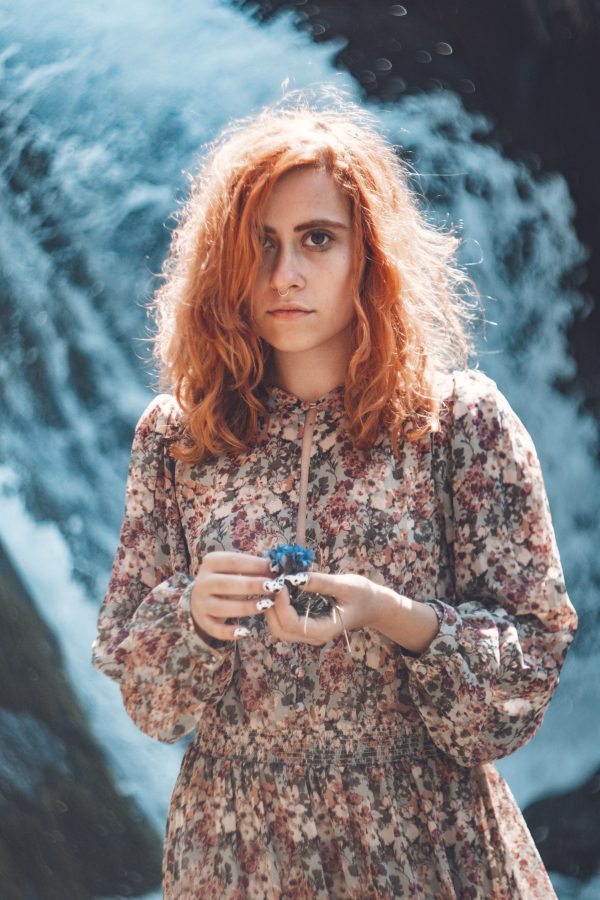 A woman with curly hair in a floral dress stands by a waterfall, holding a blue flower. Mobile 4K