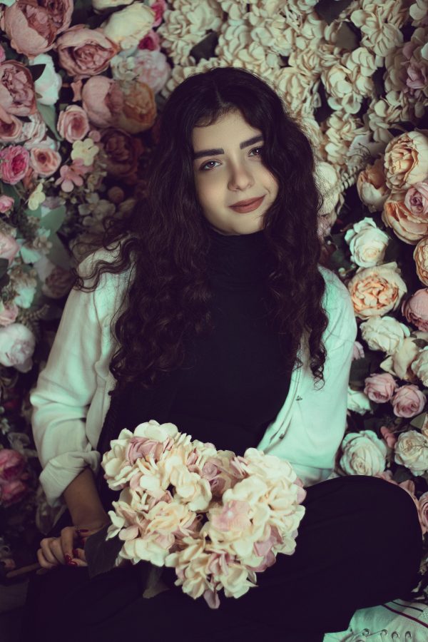 A woman with curly hair sits amidst a lush floral backdrop, holding a bouquet. Mobile 4K