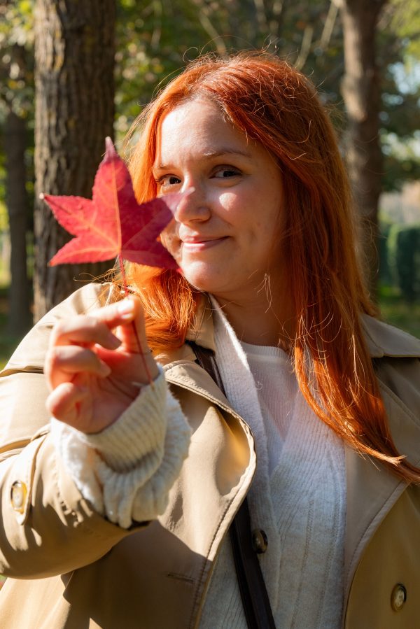 A woman with red hair holds a red leaf, smiling in a sunlit autumn park. Mobile 4K