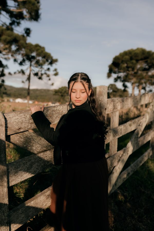 A young woman in a black dress poses gracefully beside a rustic wooden fence in a serene outdoor setting. Mobile 4K