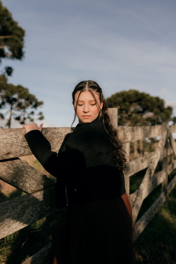 A young woman in a black jacket poses thoughtfully by a rustic wooden fence on a sunny day. Mobile 4K