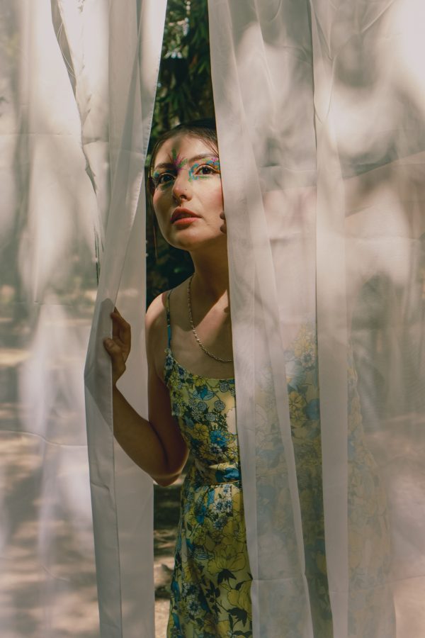 A young woman in a floral dress peers through sheer curtains surrounded by nature. Mobile 4K