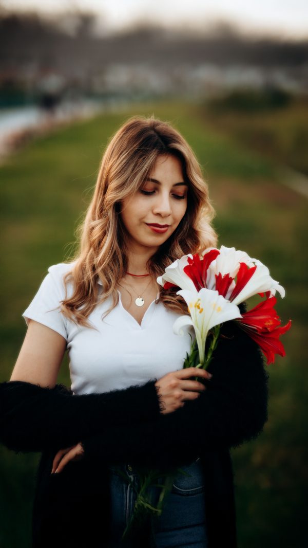 A young woman with flowing hair holds a bouquet of red and white lilies in a serene outdoor setting. Mobile 4K