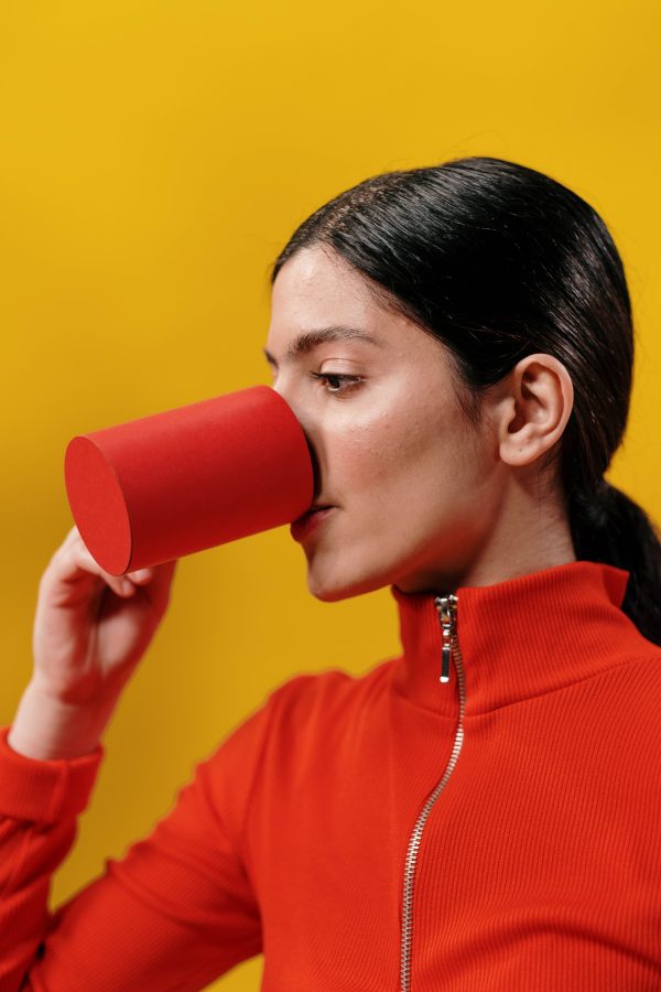 Adult woman in red drinking from a paper cup against a vibrant yellow background. Mobile 4K