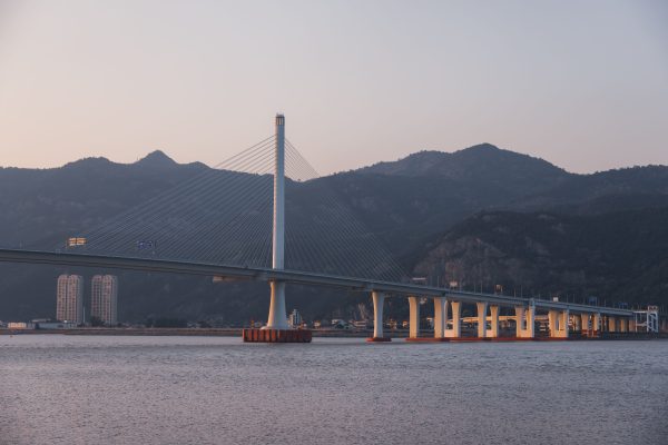 An elegant suspension bridge spans across a river at twilight with distant hills in the background. Architecture 4K