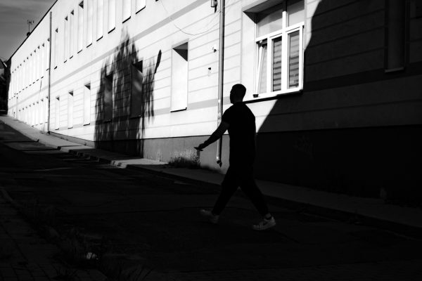 Black and white photo of a silhouette walking along a city street shadowed by a building. Dark 4K