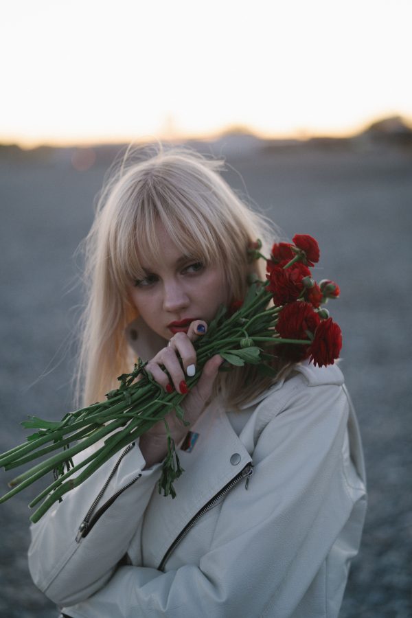 Blonde woman in white coat holding red flowers at sunset outdoors. Mobile 4K