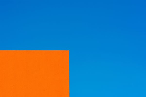 Bright orange geometric building corner set against a clear, vibrant blue sky. Minimaliste 4K