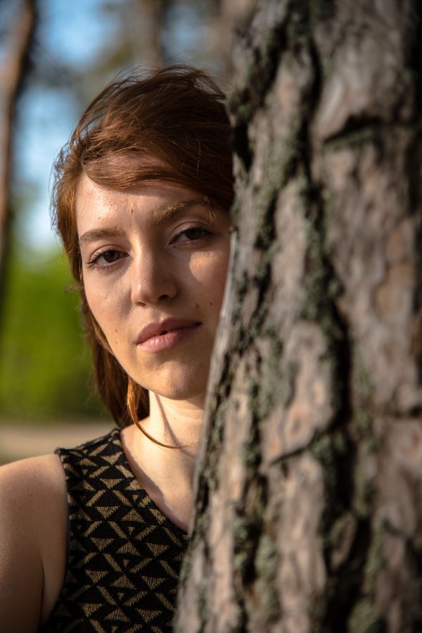 Close-up portrait of a woman with red hair leaning against a tree in a sunlit forest. Mobile 4K