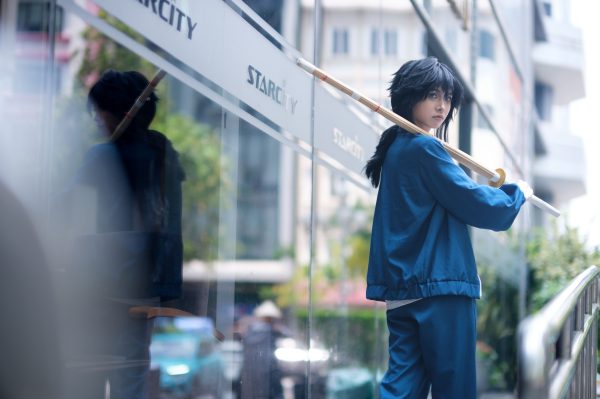 Cosplayer in blue attire holding a staff, reflecting against a glass building in an urban area. Manga 4K