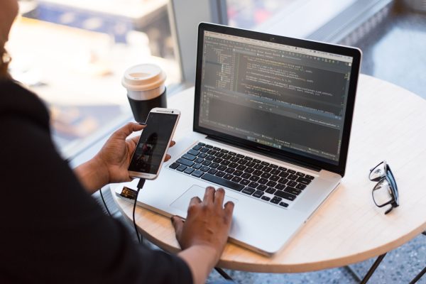 Developer working remotely, coding on a laptop with phone in hand, showcasing modern work culture. Wallpaper 4K
