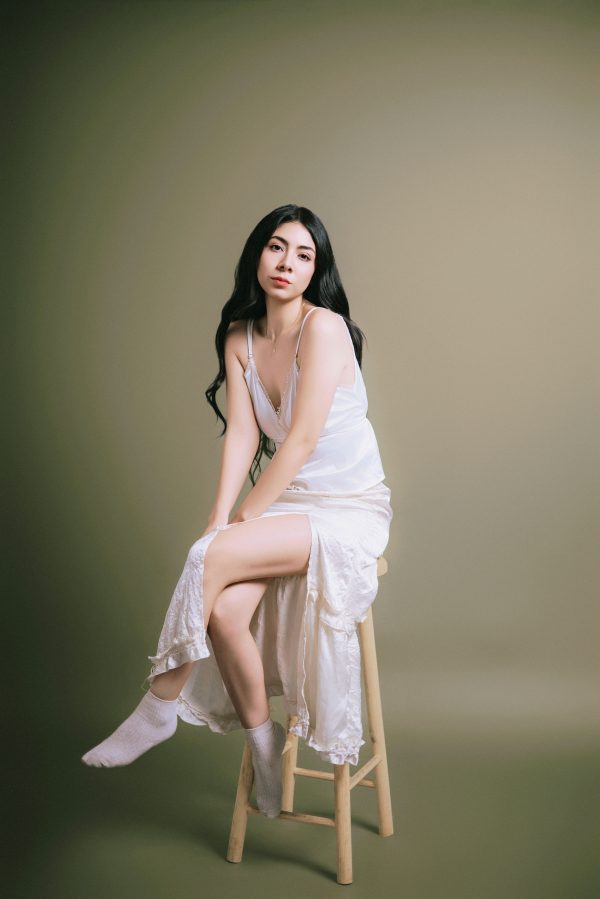 Graceful woman in a white dress sitting on a stool against a muted backdrop. Mobile 4K