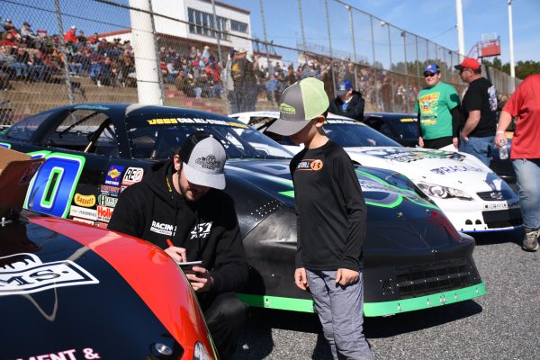 NASCAR driver signing autographs for young fan at outdoor racing event with multiple cars and spectators. Voitures 4K
