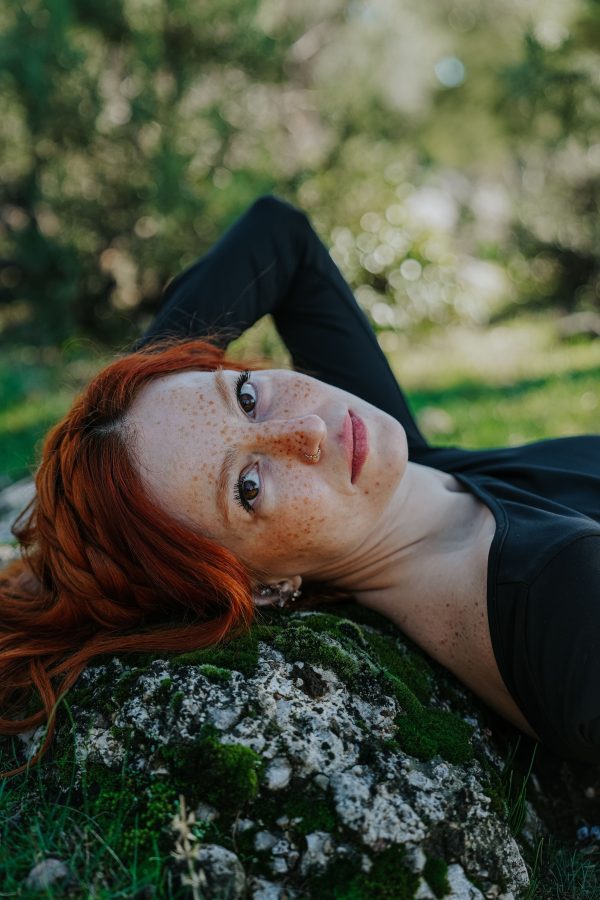 Portrait of a freckled redhead woman lying on a moss-covered rock outdoors during summer. Mobile 4K
