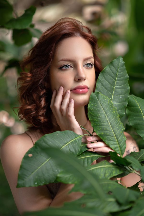 Portrait of a woman with brown hair amidst lush green leaves in natural light. Mobile 4K