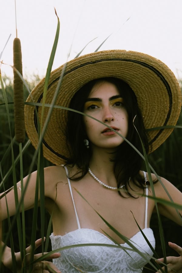 Portrait of a young woman in a white dress and straw hat standing amidst tall cattails. Summer vibe. Mobile 4K