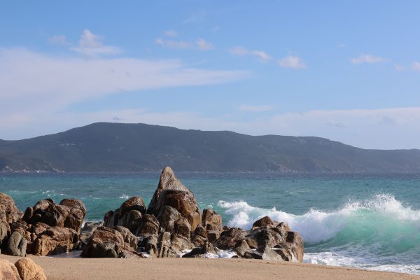 Scenic view of waves crashing on a rocky coastline with mountains in the background under a clear blue sky. Nature 4K