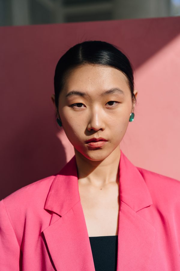 Serious portrait of a woman wearing a pink blazer with light and shadow contrast on a pink background. Mobile 4K