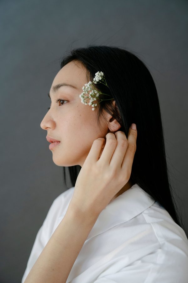 Side view of attractive Asian female in white garment and small twig in black hair standing on gray background in studio Mobile 4K