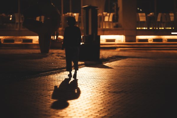 Silhouette of a woman walking on a dimly lit urban sidewalk at night, with reflections. Dark 4K