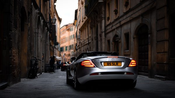 Silver luxury convertible driving through narrow, historic city street at dusk. Voitures 4K