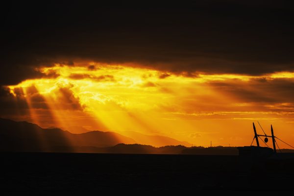 Striking sunset with sun rays piercing dramatic clouds over the ocean and a silhouetted boat. Dark 4K