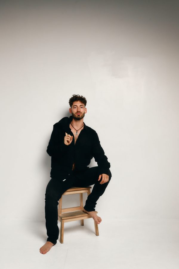 Stylish male model in black attire posing barefoot on a stool against a minimal white background. Mobile 4K