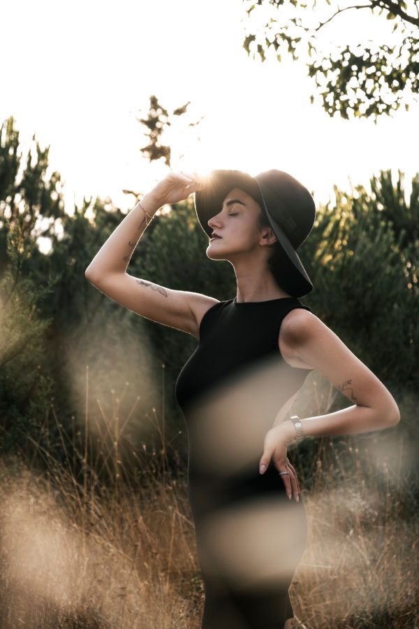 Stylish woman posing in a black dress and hat in a rural setting during the day. Mobile 4K