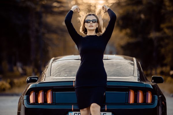 Stylish woman wearing sunglasses poses confidently next to a Ford Mustang in an outdoor setting, embodying elegance and strength. Voitures 4K